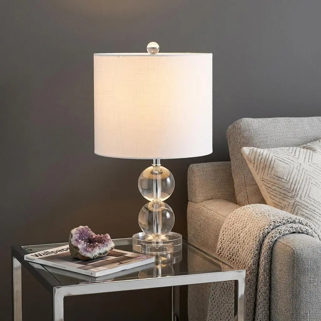 Clear acrylic table lamp with white fabric shade, modern design, and round base, styled beside a cozy gray sofa.
