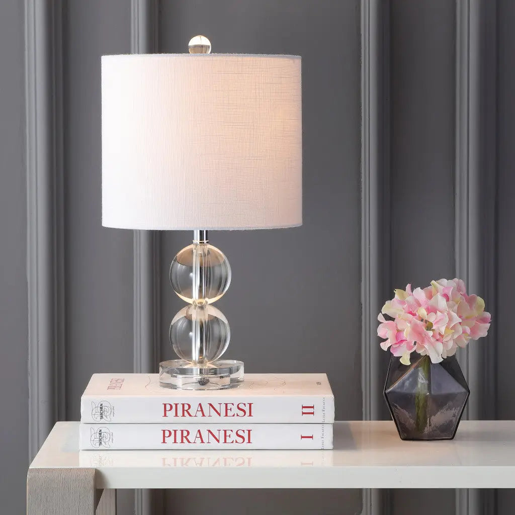 Clear glass table lamp with white shade, modern design, placed on stacked books beside a geometric vase of pink flowers.