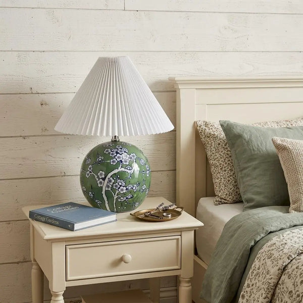 Green floral ceramic table lamp with white pleated shade on a beige nightstand in a cozy bedroom setting.