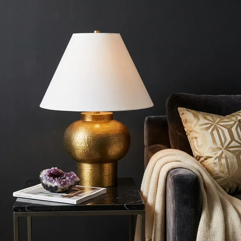 Gold hammered table lamp with white shade, modern design, placed on a black marble table beside a plush gray sofa.