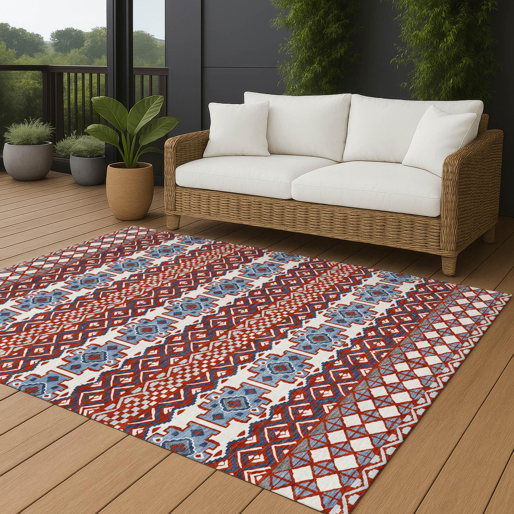 Red and blue patterned outdoor rug on a wooden deck with a wicker sofa and greenery in the background.