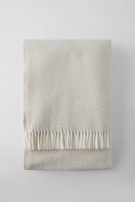 Light beige woven blanket with fringe edges, made of soft cotton for a cozy, stylish décor accent.