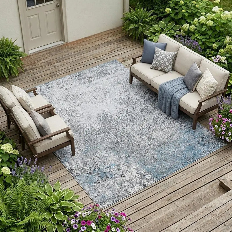 Blue and gray patterned outdoor rug on a wooden patio, surrounded by lush greenery and colorful flowers.